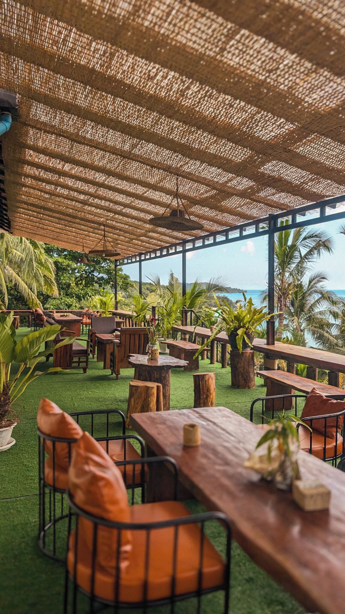An outdoor dining area with wooden tables and chairs, surrounded by greenery and overlooking a scenic view.