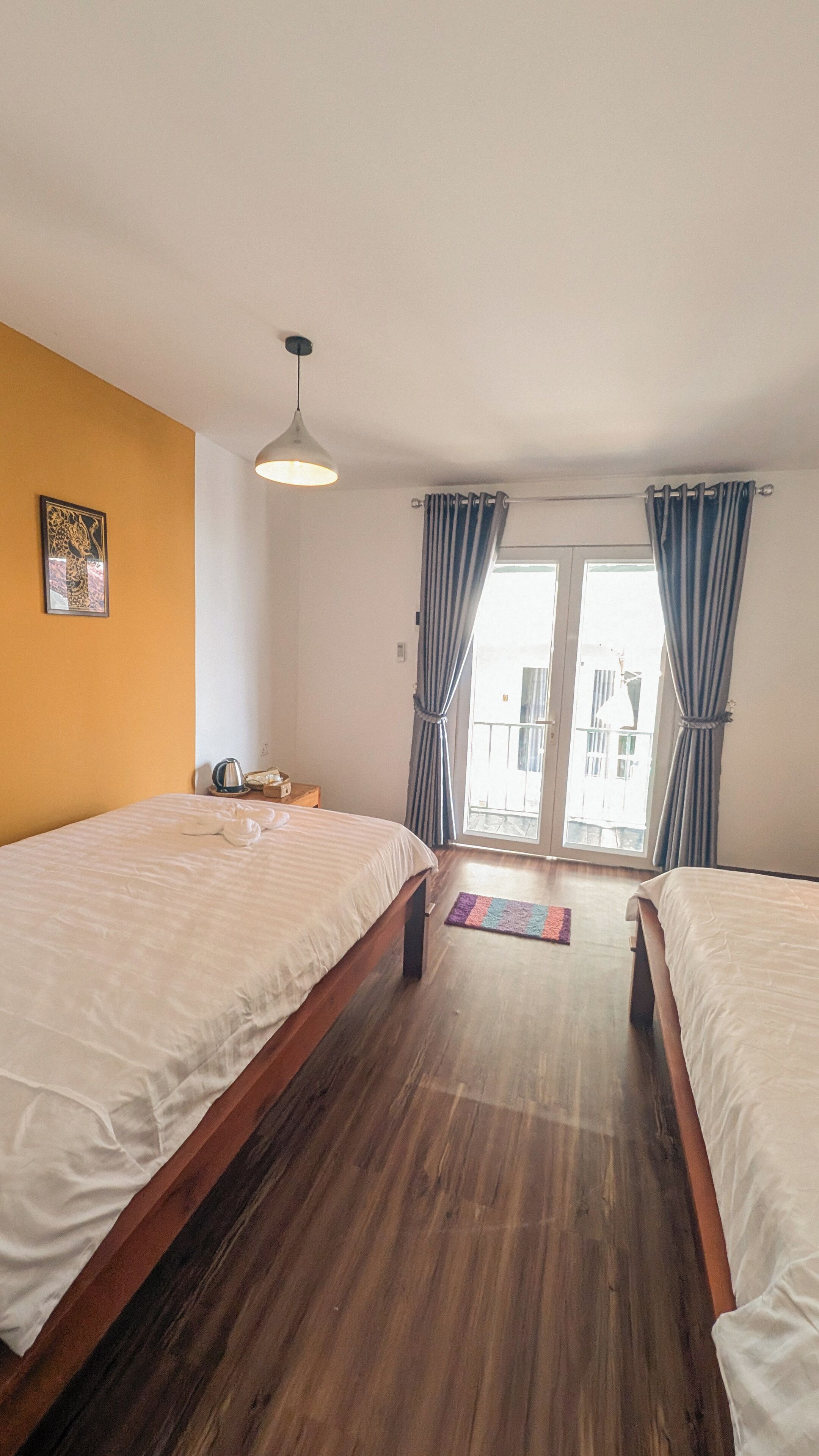 A room with two beds, a ceiling light, a window with curtains, and a door leading to an outdoor balcony.