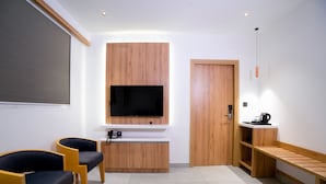 A modern room with a flat-screen TV mounted on a wooden panel, two chairs, a coffee machine, and a wooden bench. - AJ Palace (Ambalapuzha)