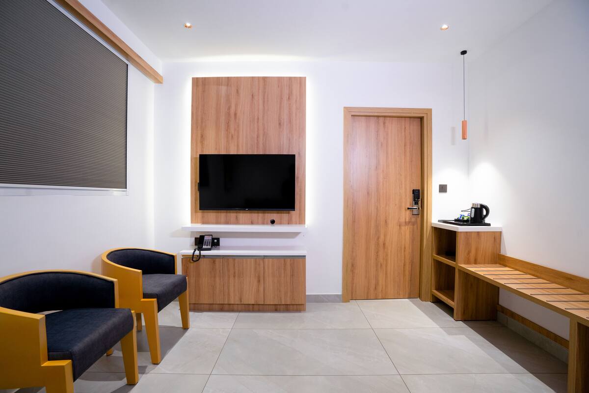 A modern room with a flat-screen TV mounted on a wooden panel, two chairs, a coffee machine, and a wooden bench.
