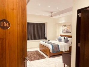 A bedroom with a wooden door marked "104", a bed with a blue and white bedspread, a window with curtains, and a painting on the wall.