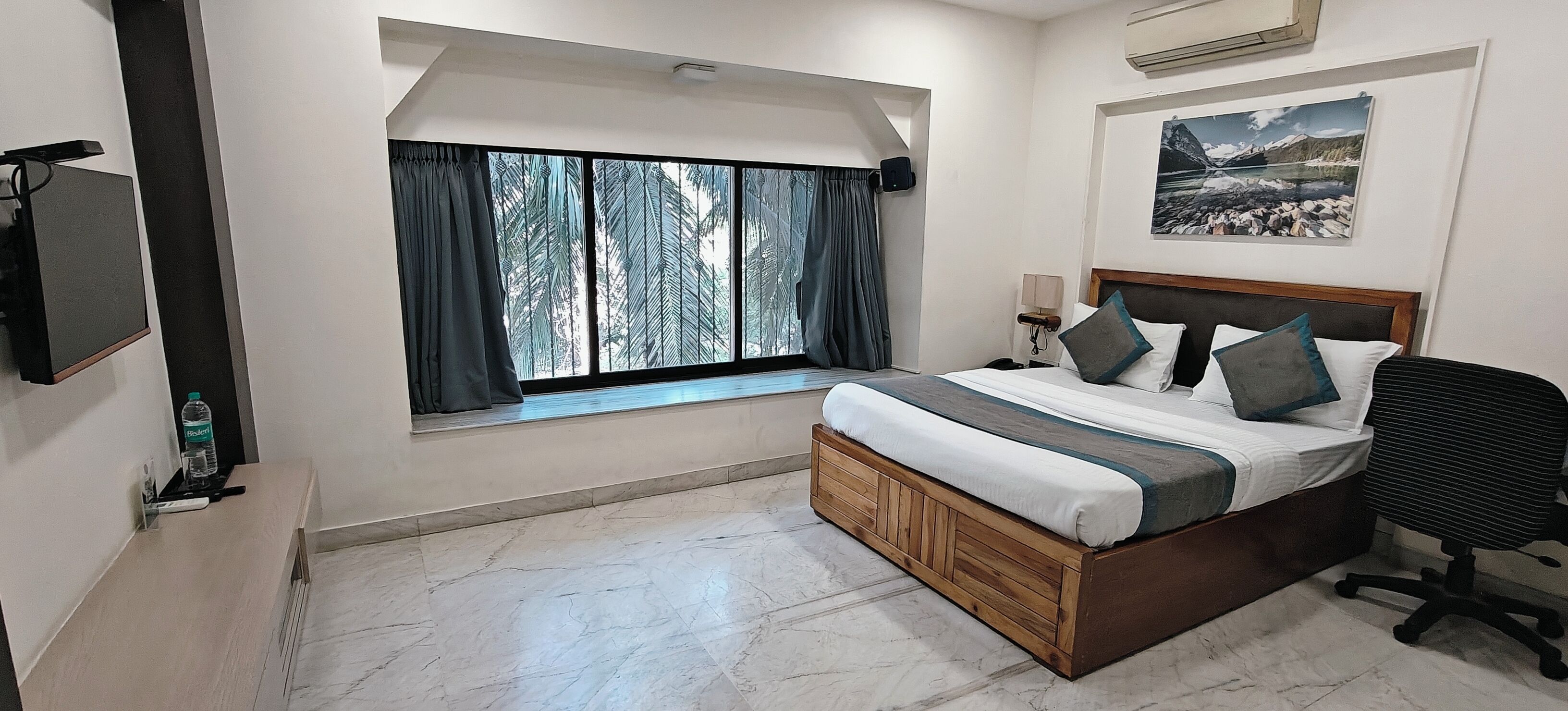 A bedroom with a bed, a desk, a chair, a window with curtains, a wall-mounted TV, and a painting.