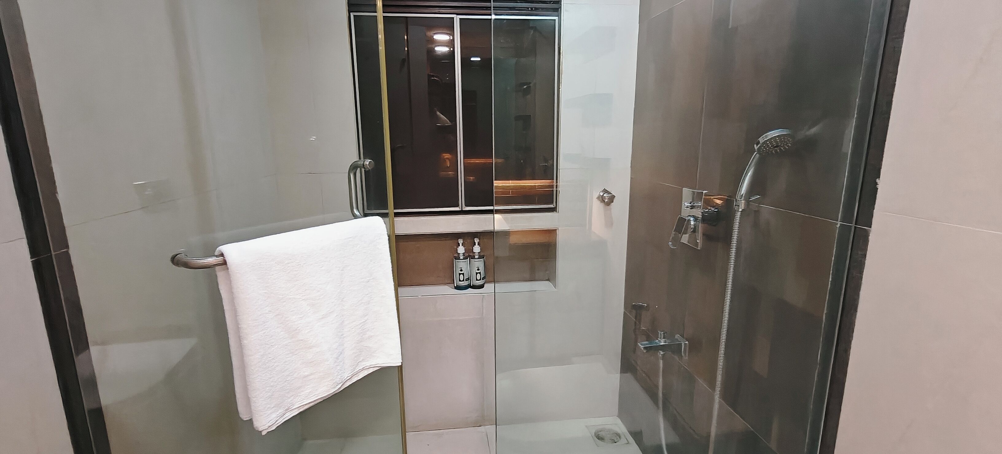 A modern bathroom with a glass-enclosed shower, a white towel hanging on a metal towel rack, and a small shelf with two bottles.