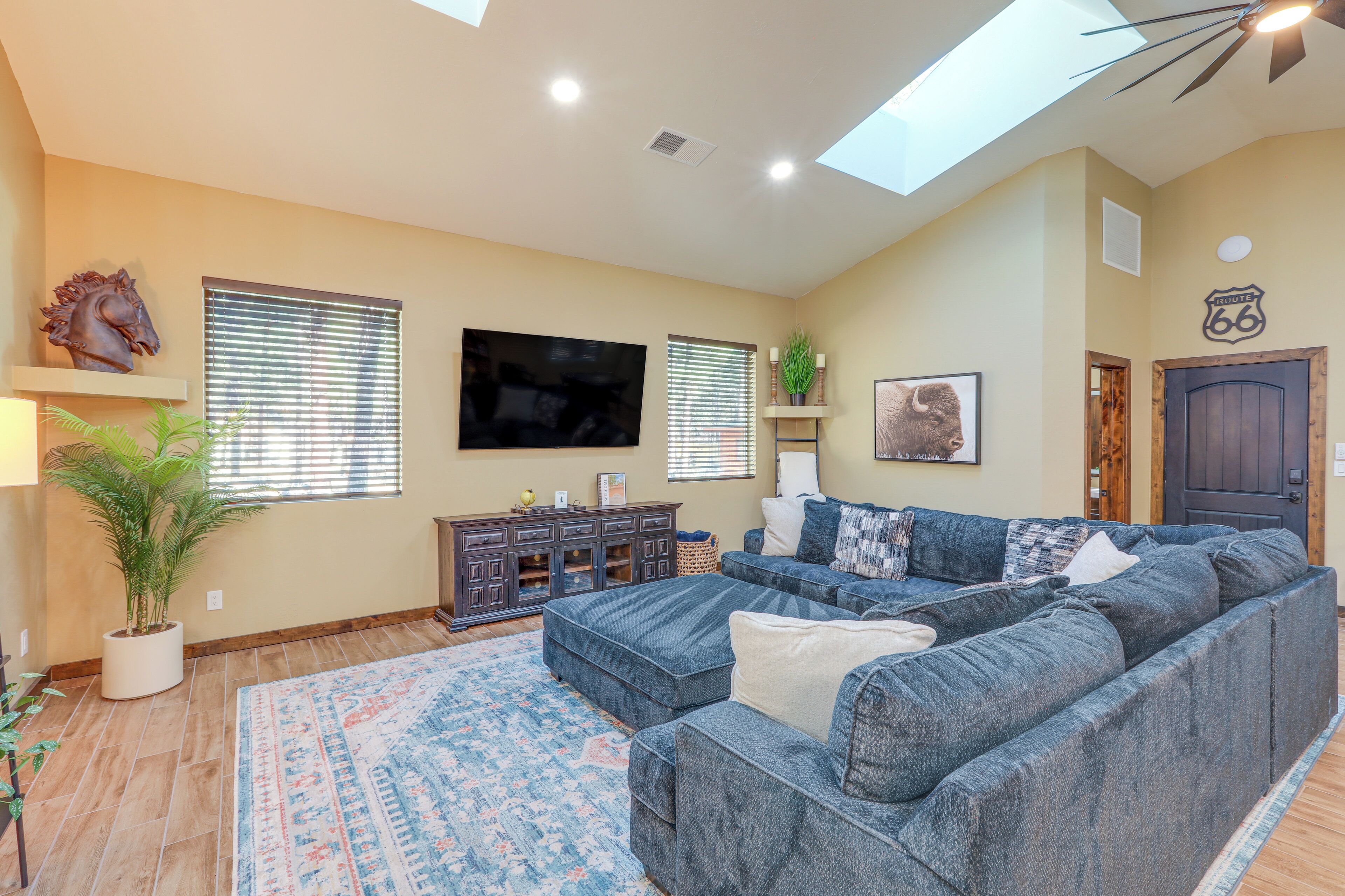 A spacious living room with a sectional sofa, a flat-screen TV, and a large window with blinds.