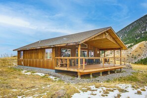 Una cabaña de madera con terraza, amplios ventanales y montañas al fondo.