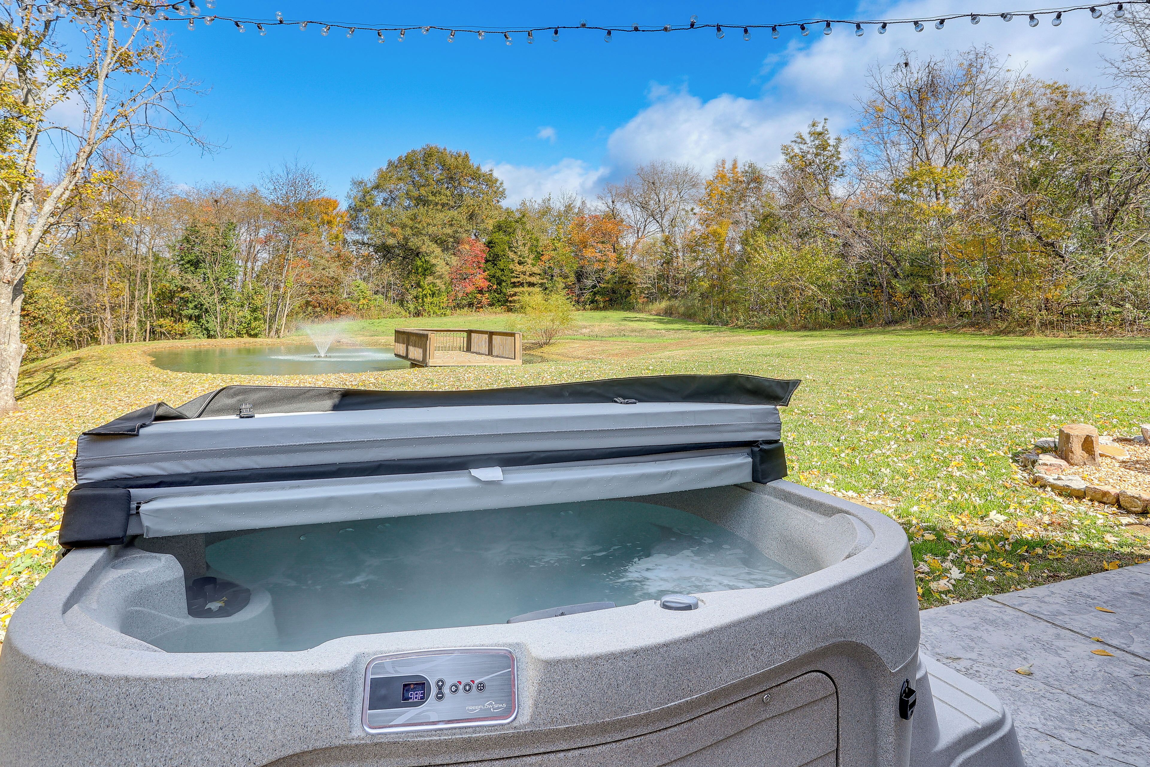 A hot tub in a backyard with a wooden deck, a pond, and trees in the background.