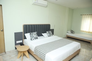 A hotel room with a large bed, a bedside table, a small stool, a window with curtains, and a wall-mounted air conditioner. - Hotel Rangoli (Udaipur)