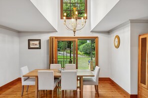 Una sala da pranzo con tavolo in legno e sedie bianche, un lampadario, un quadro incorniciato e vista su un giardino attraverso una porta di vetro.