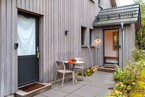 Une maison moderne à l’extérieur gris, avec une porte d’entrée sombre, un petit patio doté d’une table ronde et de deux chaises, et un jardin agrémenté de plantes et d’un banc en bois.