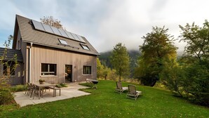 A modern house with a sloped roof, solar panels, a patio with a table and chairs, and a well-maintained lawn.