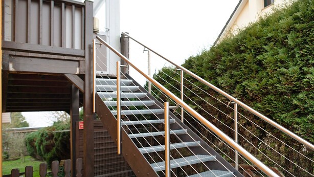 A metal staircase with a handrail leading to a covered area, surrounded by greenery and a building.