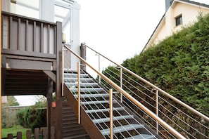 Una escalera metálica con barandilla que conduce a una zona cubierta, rodeada de vegetación y un edificio.