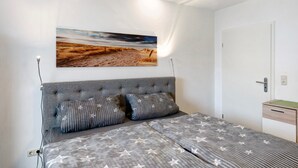 A bedroom with a bed, a headboard, two pillows, a nightstand, a wall-mounted picture, and a door.