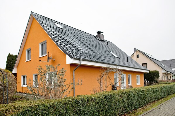 Une maison moderne à deux étages, dotée d’un toit en pente, d’une cheminée bien visible et d’une haie bien entretenue à l’avant.