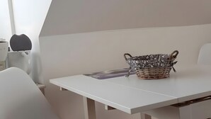 Une table blanche avec un panier tressé, une chaise blanche et un mur blanc orné d’un luminaire aux motifs géométriques.