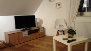Un salon moderne avec un téléviseur à écran plat, un meuble en bois, une chaise blanche, une petite table avec une télécommande et une plante en pot.
