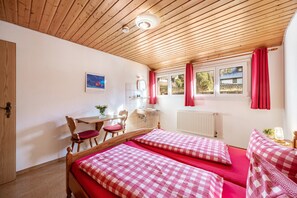 A room with two beds, a wooden desk, a chair, a window with curtains, and a radiator.