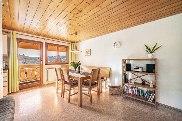 A wooden interior with a dining table, chairs, a bookshelf, and a view through a window.