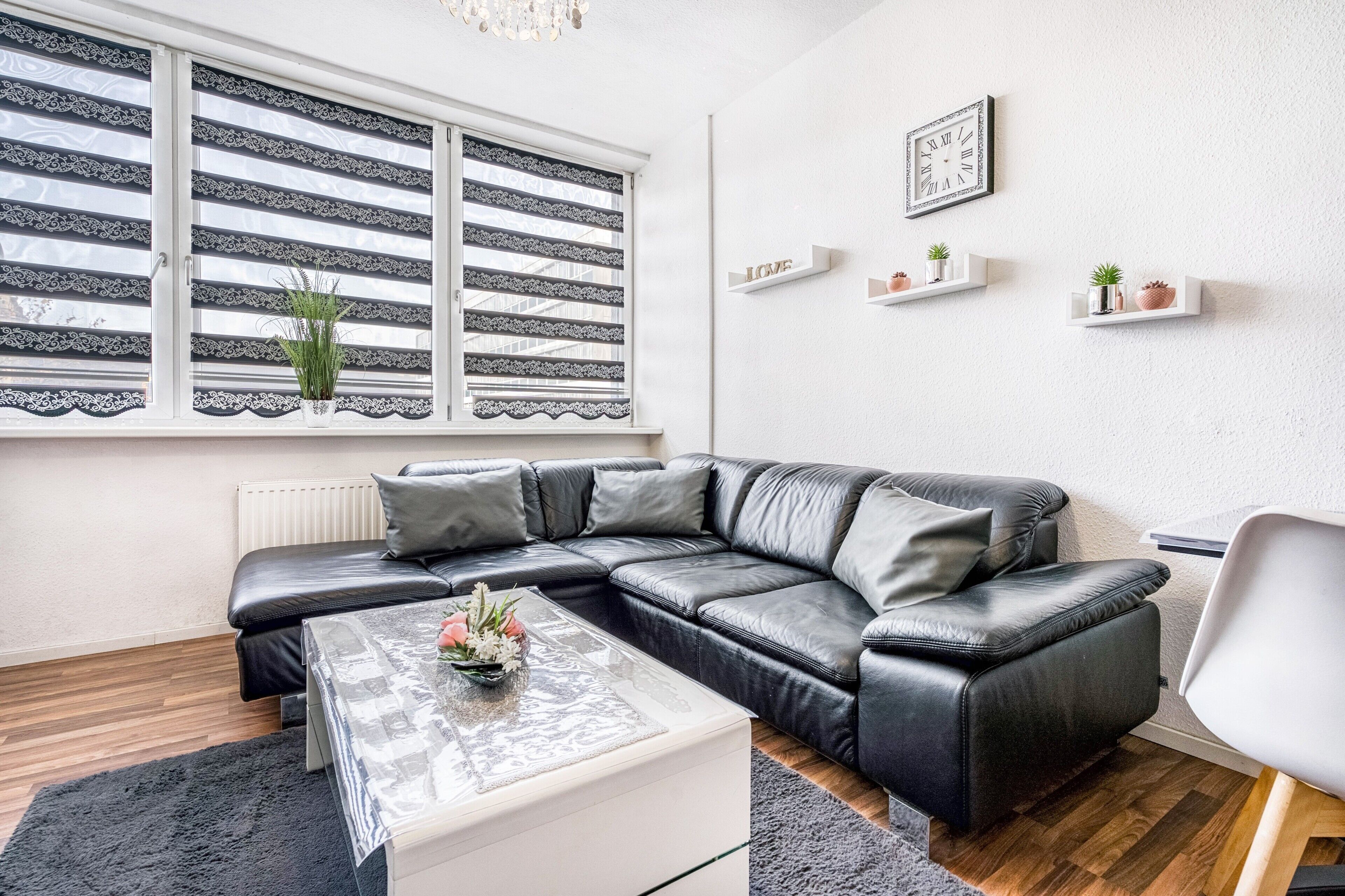 A modern living room with a black leather sectional sofa, a glass coffee table, and a white radiator. There are decorative shelves with plants and a framed clock on the wall.