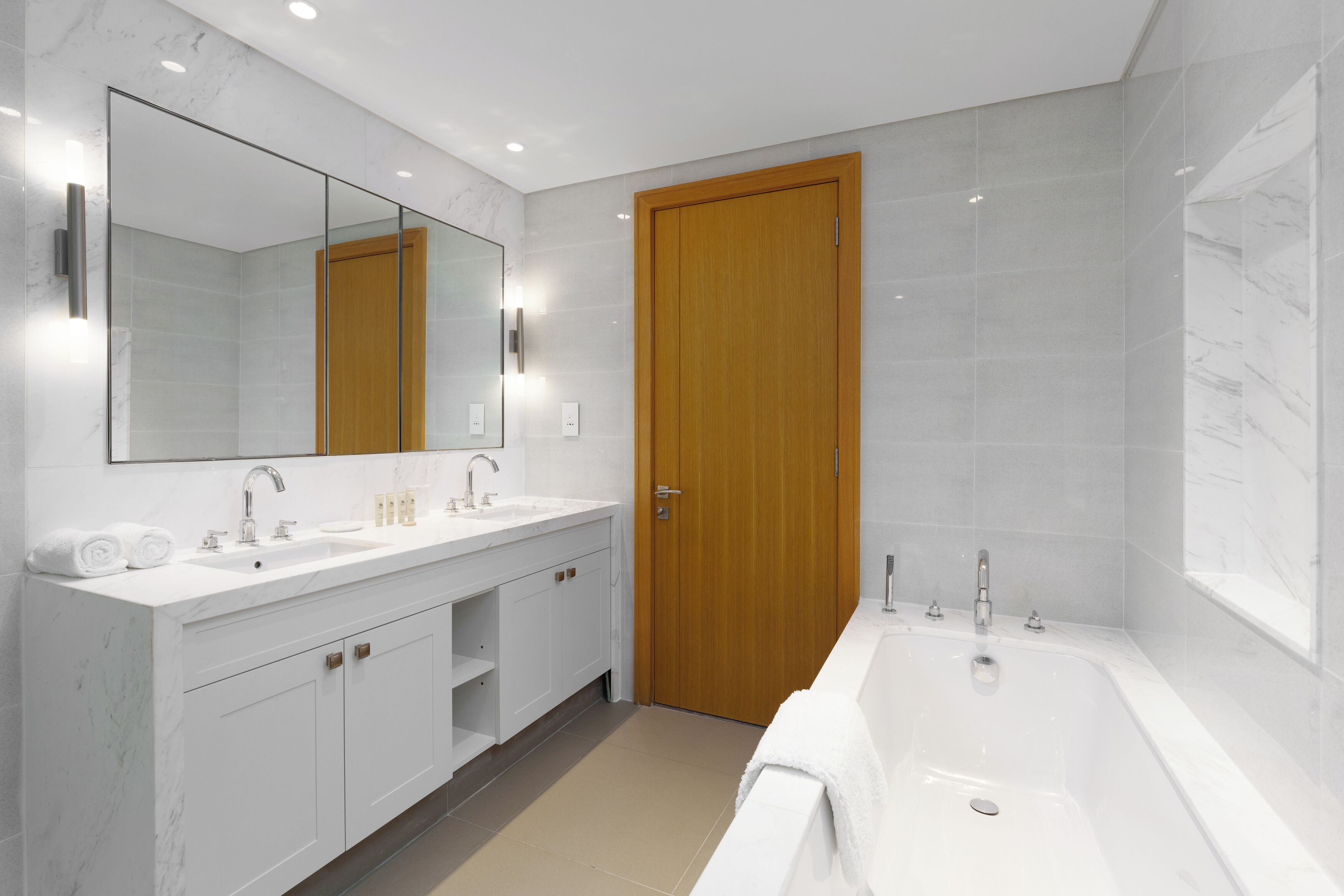 Een moderne badkamer met een grote spiegel, twee wastafels, een badkuip en een houten deur.