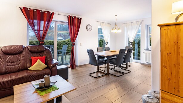 En moderne stue med en lædersofa, spisebord og stole, store vinduer med gardiner, samt et træskab.