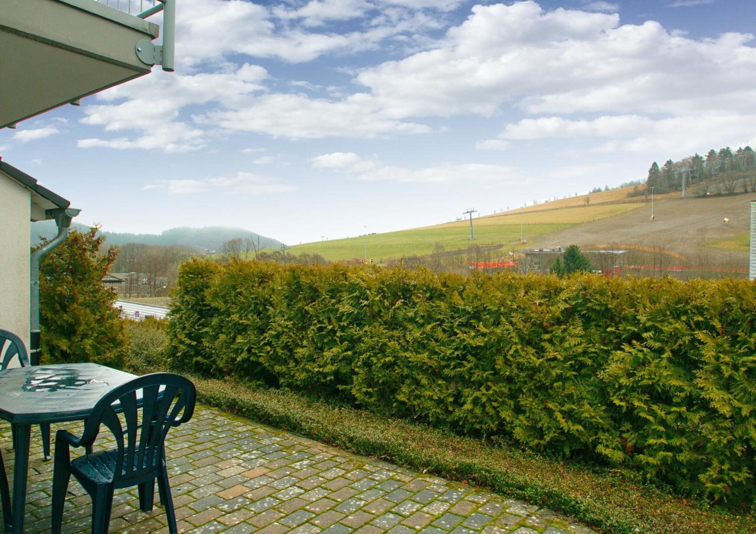 A patio with a table and chairs, a hedge, and a view of a green landscape with hills and a clear sky.