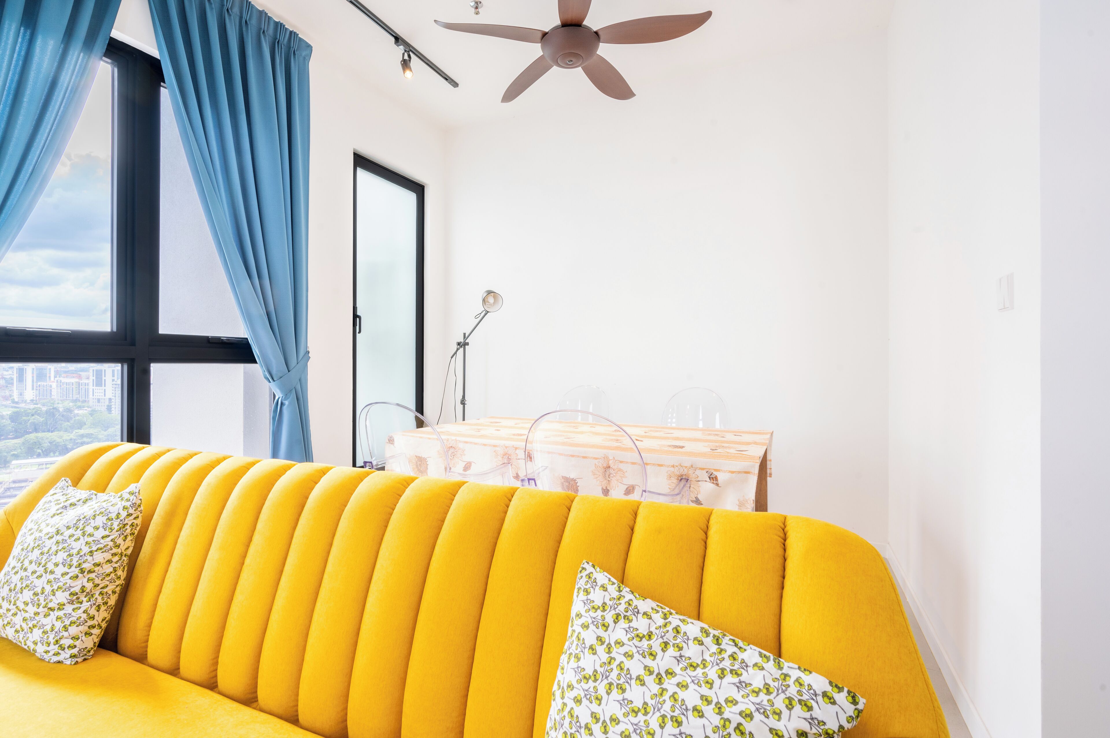 A bright yellow sofa with floral cushions, a ceiling fan, a window with blue curtains, and a desk with transparent storage containers.