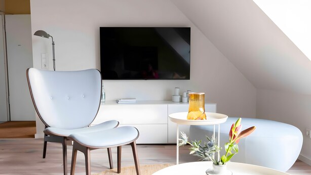 Ein modernes Wohnzimmer mit Fernseher, einem hellblauen Sofa, einem weißen runden Tisch mit einer Vase und einem grauen Sessel.