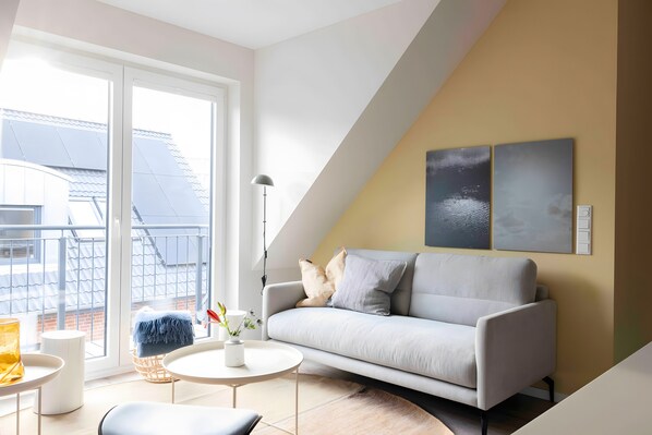 Un salon moderne avec un canapé, une table basse et un lampadaire. Une grande fenêtre offre une vue sur un immeuble.