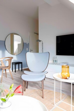 Una habitación de hotel moderna con un espejo redondo, una silla blanca, una mesita con una vela amarilla y un televisor de pantalla plana.