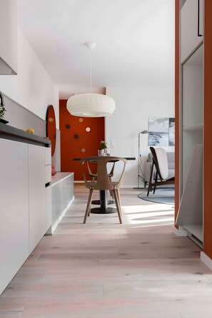 A modern kitchen with a dining area, featuring a wooden table and chairs, a pendant light, and a view of the living room.