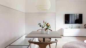 Un espace repas moderne avec une table en verre, des chaises en bois, un luminaire suspendu blanc et un téléviseur à écran plat fixé au mur.