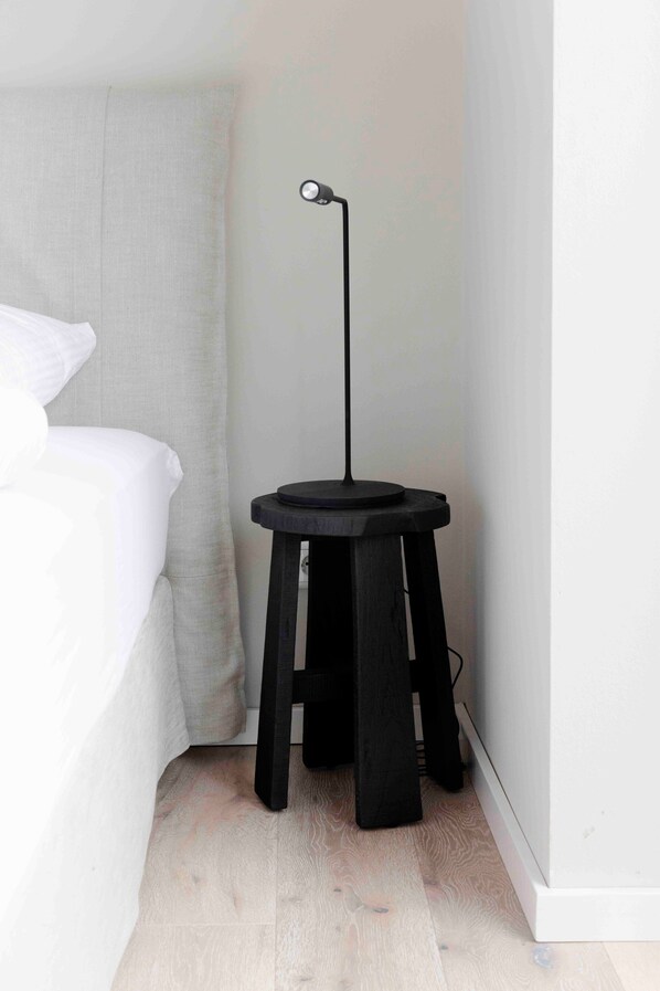 Dormitorio en esquina con cama, un taburete negro, una lámpara de pie y una pared blanca.