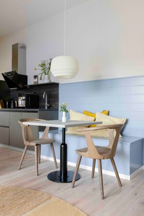 Una cocina moderna con comedor, que presenta una mesa blanca, dos sillas de madera, una lámpara colgante y un banco empotrado con cojines amarillos.