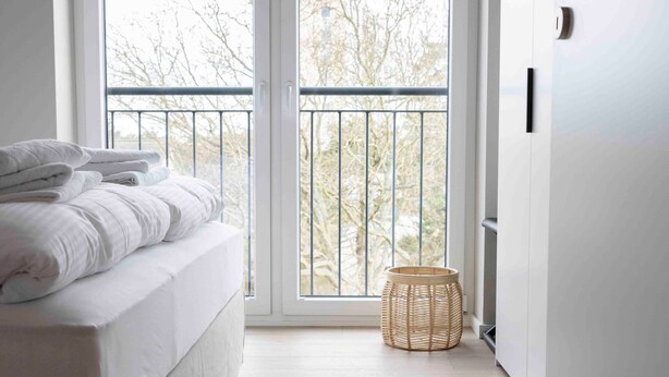 A bedroom with a bed, a window, a door, and a small wicker basket. - Apartment 'Ocean Star' with Balcony and Wi-Fi (Sankt Peter-Ording)