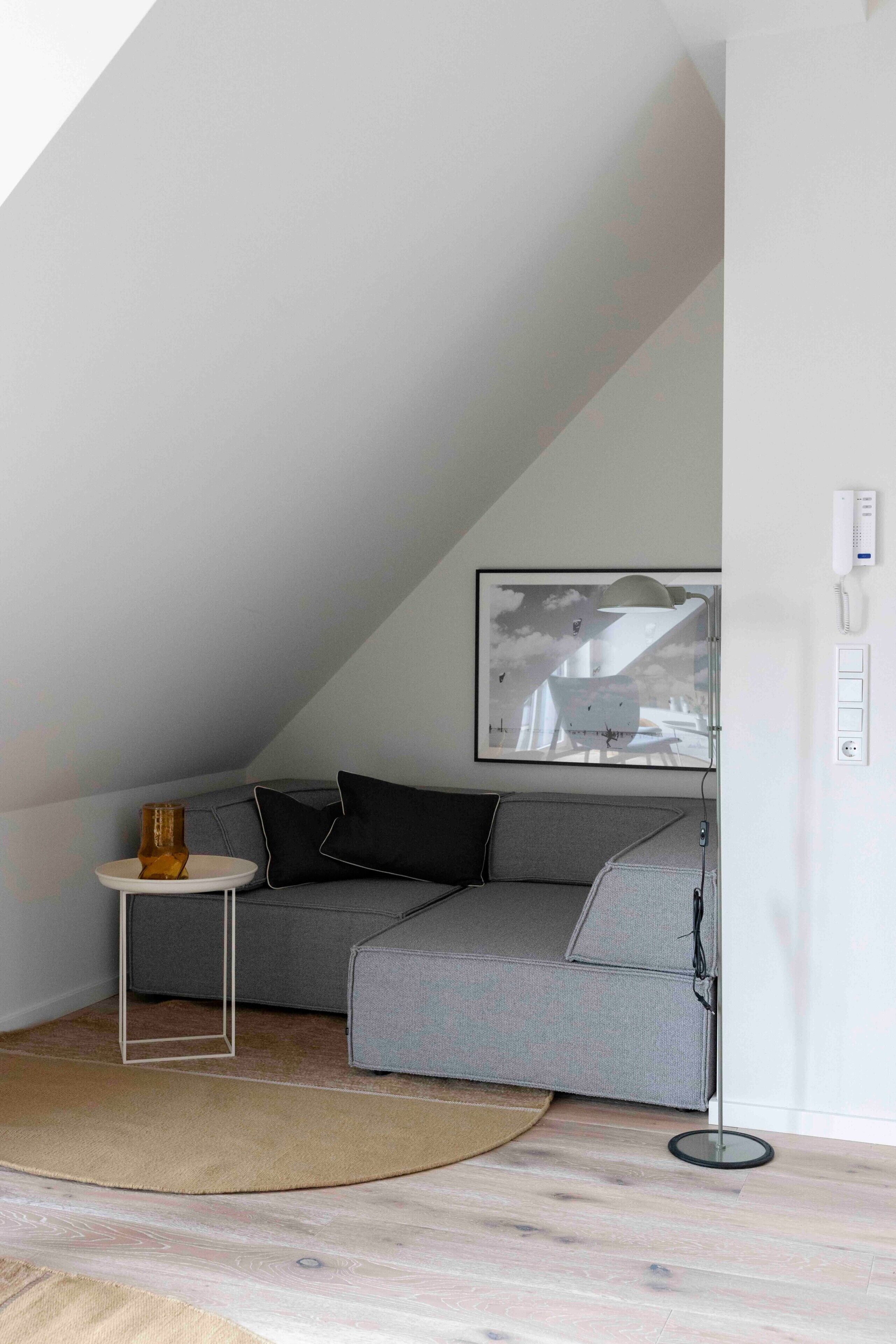 Ett modernt sovrum i loftstil med en grå, modulbaserad soffa, ett litet runt bord och en inramad tavla på väggen.