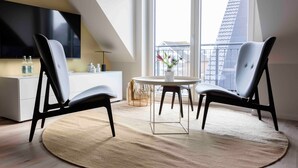 Una sala de estar moderna con televisor, dos sillones, una mesa redonda y vistas de la ciudad a través de amplios ventanales.