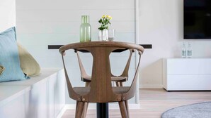 A modern dining area with a round table, wooden chairs, a hanging light fixture, a sofa with cushions, and a TV on a white cabinet. - Apartment 'Sunseeker' with Balcony and Wi-Fi (Sankt Peter-Ording)