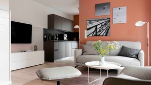 A modern living room with a sofa, coffee table, and ottoman, a kitchen area, and a flat-screen TV. - Apartment 'Invictus' with Private Terrace and Wi-Fi (Sankt Peter-Ording)