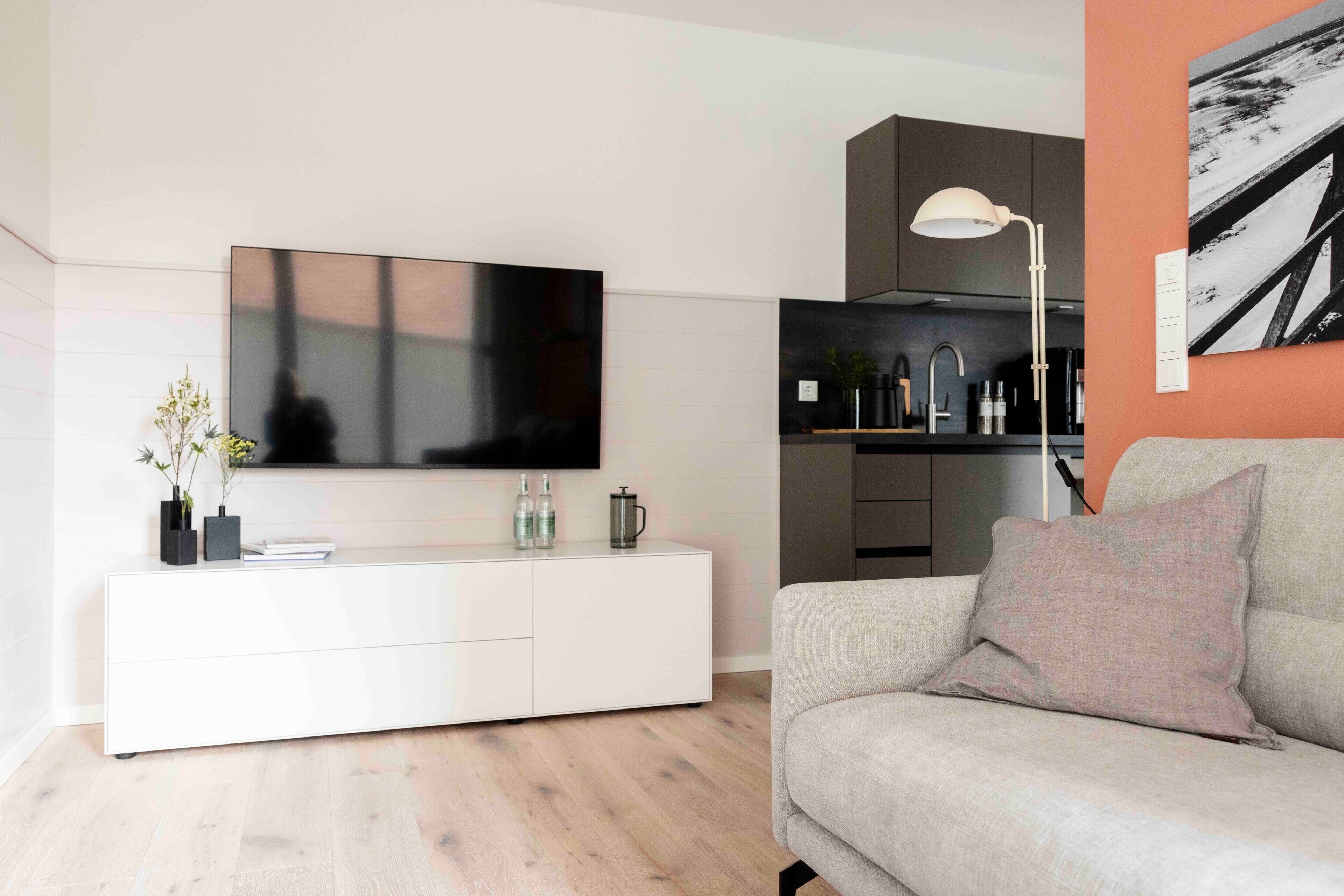 A modern living room with a flat-screen TV, white cabinetry, a grey sofa, and a kitchen area in the background.
