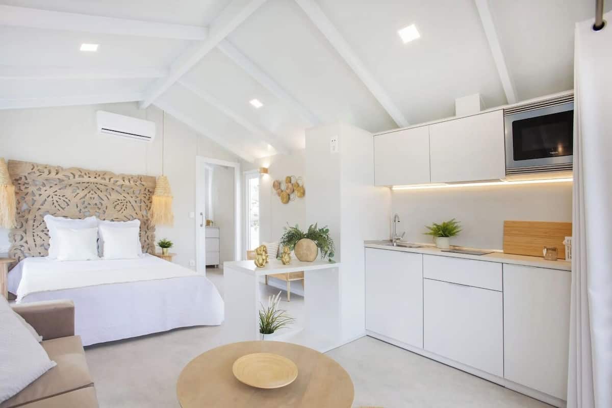 A modern hotel room with a bed, a sofa, a small coffee table, a kitchen area with a TV, and a dining table.