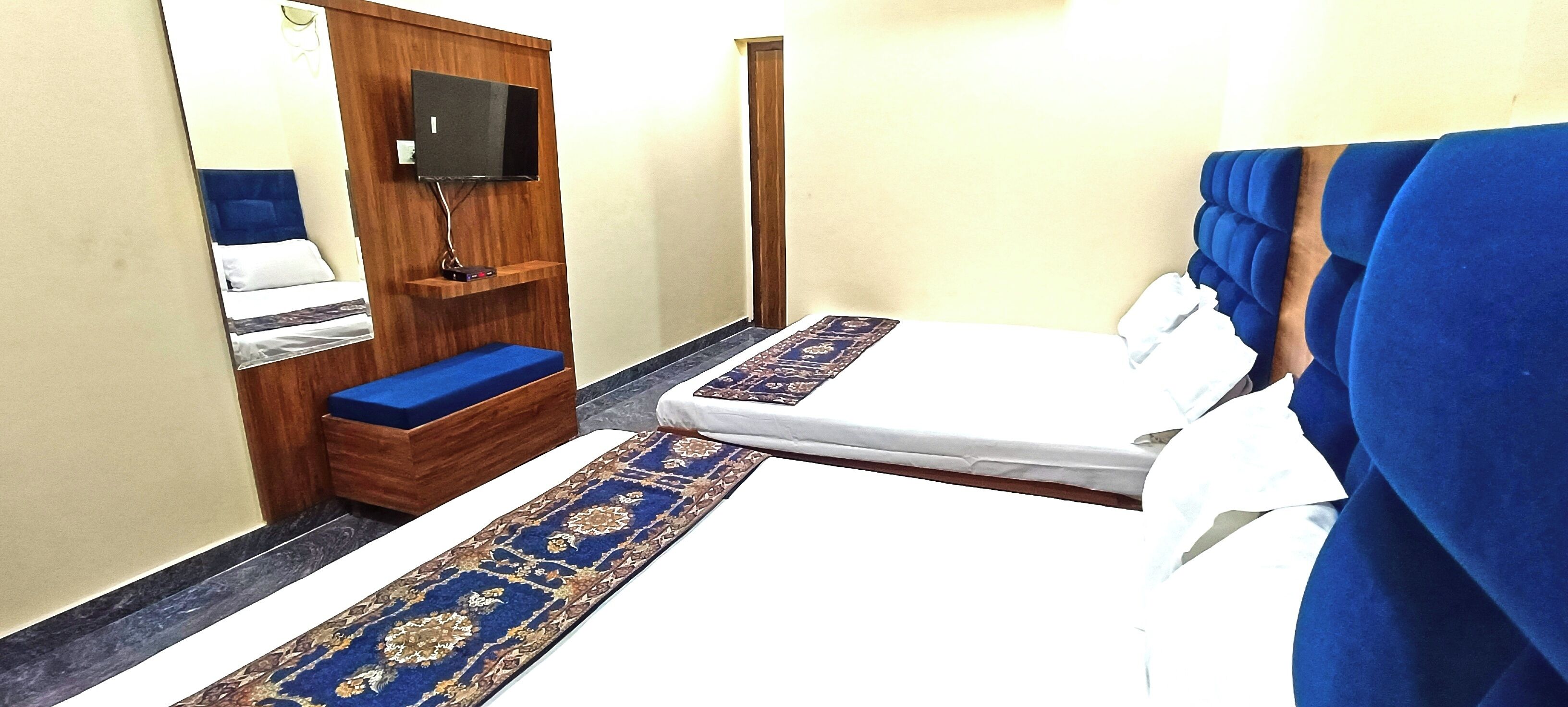 A hotel room with a bed, a bench, a TV mounted on the wall, and a carpet runner.