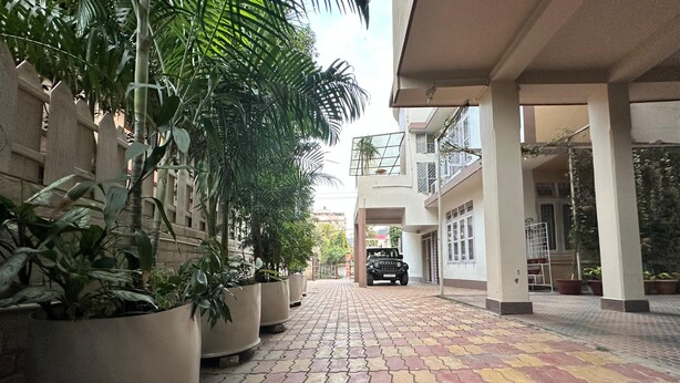 A paved walkway with potted plants on the left, a black vehicle parked at the end, and a building with balconies and large windows on the right. - The Dew Drop Hotel (Guwahati)