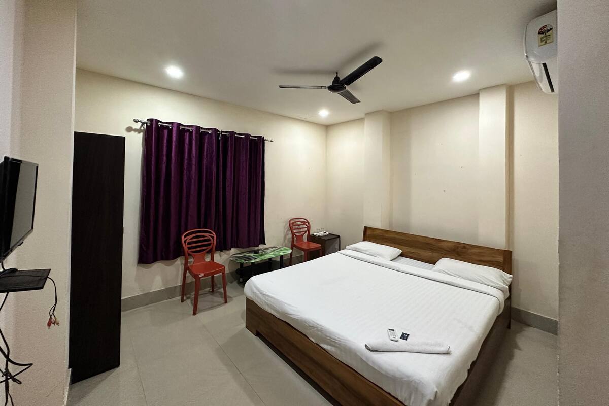 Ein Hotelzimmer mit einem Bett, zwei Stühlen, einem Schreibtisch, einem Fernseher, einem Deckenventilator und einem Fenster mit violetten Vorhängen.