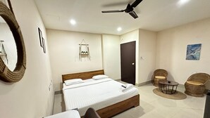 A hotel room with a bed, a desk, two wicker chairs, a ceiling fan, and a door. - The Dew Drop Hotel (Guwahati)