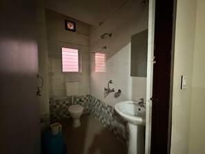 Een kleine badkamer met toilet, bidet en wastafel. Er zijn twee ramen met zonwering en de muren zijn betegeld.