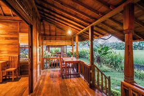 Un porche con suelo de madera, zona de comedor, barandillas de madera y vistas a un jardín exuberante.