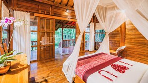 Une cabane en bois avec un lit à baldaquin, une orchidée en pot et une vue sur une terrasse en bois et la verdure environnante.