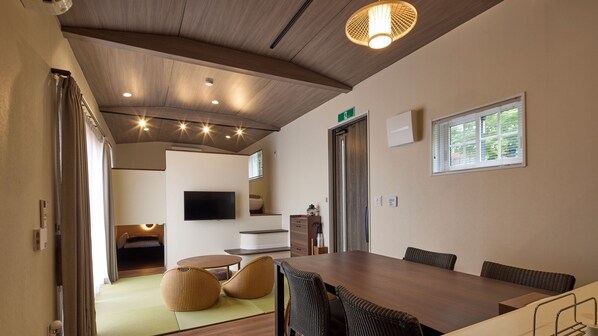 Un interior moderno con techo de madera, una zona de comedor con una mesa grande y sillas, un televisor montado en la pared y una ventana con persianas.
