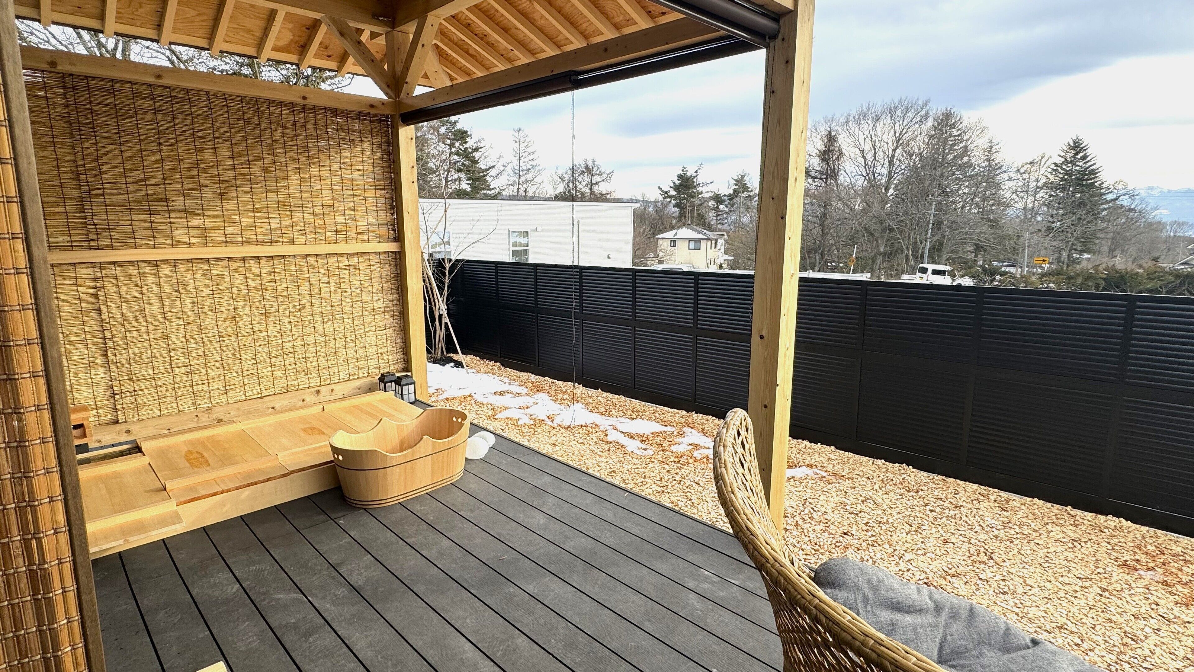 A covered outdoor area with a wooden deck, a bamboo wall, a black privacy fence, and a wicker chair.